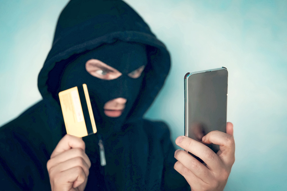 Close up of a man in robbery mask and hood holding the credit card and looking at the smartphone screen. Male criminal arranges a financial affair with mobile and credit card. Network fraud dangers.